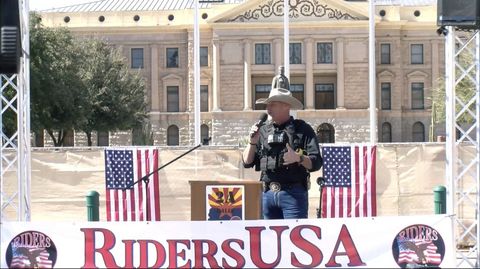 Sheriff Mark Lamb, wearing a big white cowboy hat and his usual black Sheriff vest stand on stage in front of the Phoenix Capitol. Two American flags are pinned on the fence behind him. A large white banner with two logos for Riders USA reads “Riders USA Celebrate & Protect the 2nd Amendment.’ Video camera and sound speakers can be seen set up around the stage Lamb is standing on.
