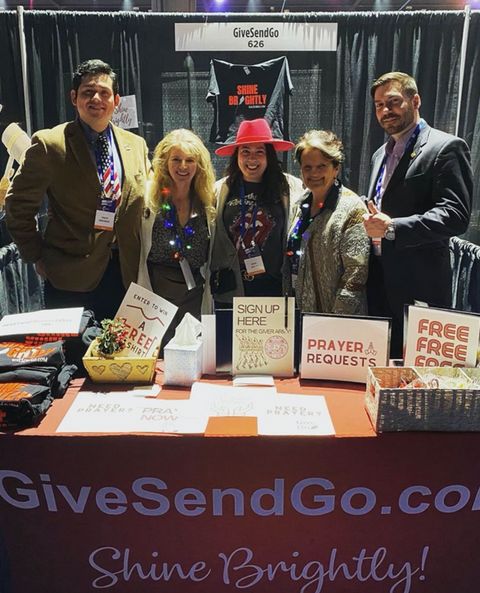 Dio Brando on left and Mark Ivanyo on right of Republicans for National Renewal pose with a group of women running the Give Send Go booth inside the exhibitor hall of AmericaFest