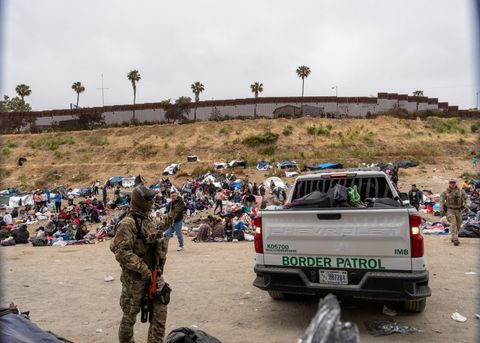 a border patrol agent in heavy camo gear with a rifle stands next to a border patrol truck in front of the camp