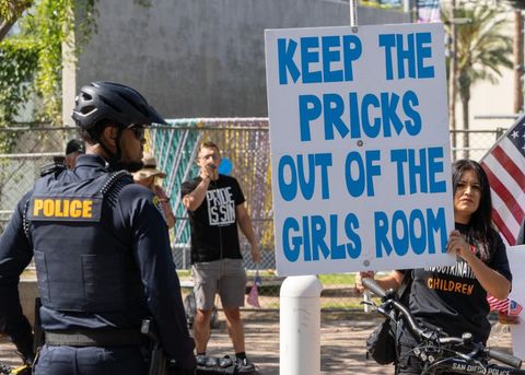 A police officer stands with their back to the camera. Beyond them is a protester with a sign that reads, "keep the pricks out of the girls room." Further back, a protester is speaking into a microphone while wearing a shirt that reads, "pride is sin."