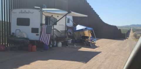 a white StarCraft RV is parked directly against the border wall, with a camp set up including a pop-up shaser, gas cans, a cooker, and chairs. An American flag and Q flag are hung on the camper.