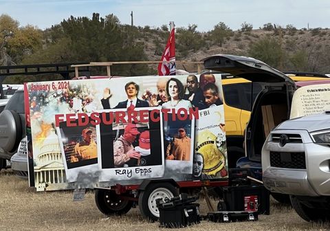 a massive, well printed sign attached on wheels to the back of a van. It reads “January 6, 2021 FEDSURRECTION” in red letters. The collage on the sign shows the Capitol riot in one corner, the Capitol in another, another side shows photos of police officers and other witnesses to the January 6th committee, Joe Biden, Nancy Peloso, Obama, and Stewart Rhodes are also blended in. Layered on top of the collage are three photos of Ray Epps on January 5 edited to look like classic polaroids. The vehicle sits in a parking lot around other cars. The cactus-filled desert and blue sky are seen in the background.