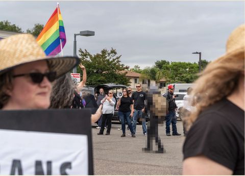 Pride attendees are visible but out of focus in the frame as they pass by the camera. In the distance, a group of far-right hecklers can be seen shouting at the march with a megaphone and filming attendees.