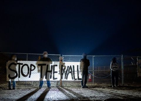 Four protesters stand in frame, holding up a banner that reads "STOP THE WALL." They are backlit by car headlights before dawn. Behind the fence, a private security employee is walking to the right.