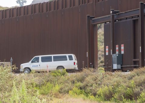 A white van with the Spencer Construction logo drives away from the border wall. The access gate is partially open.