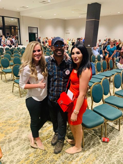 Enrique Tarrio, center, smiles indoors wearing a baseball cap and sunglasses while wrapping his arms around and posing with Lauren Wtizke, left, and Laura Loomer, right.