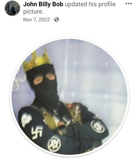 A man in a balaclava with a crown and his arms crossed. his jacket has a swastika and other nazi signage on it