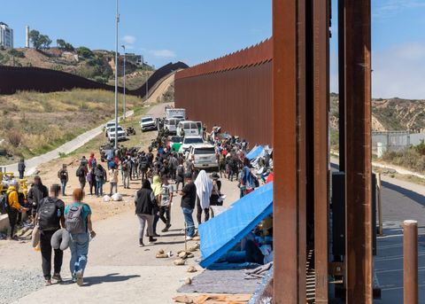 Hundreds of migrants, some standing and some in tents, are held between two massive walls. One wall terminates just in front of the camera, as the point of view is angled through a temporarily opened gate. The walls extend into the distance. Numerous border patrol vehicles and an ambulance are visible in the distance.
