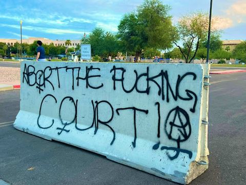 a concrete slab blocking off parking near the Capitol defaced with “ABORT THE FUCKING COURT” in black graffiti. The “O” in “abort” and “court” is the female symbol. At the end is the female symbol again but with the anarchist “A” inside.