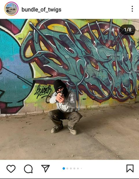instagram post from bundle of twigs showing him squatting holding a gun with his finger on the trigger in front of some graffiti 
