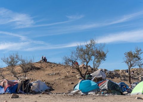 There are tents in the foreground, but distant from the camera. This wide shot contains a hilltop, and at its crest sit two asylum seekers. They are wearing sweaters. The trees are hardy desert fauna with hardly any leaves. The sky is a wide open blue with wispy streaks of clouds, and it's mid-day.