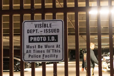 a man in a hoodie and shorts walks away from the camera. In the foreground, the bars of the police department’s parking garage gate partially obscures the interior and the walking man. In the foreground, a sign reads 'visible department-issued photo ID must be worn at all times in this police facility.'