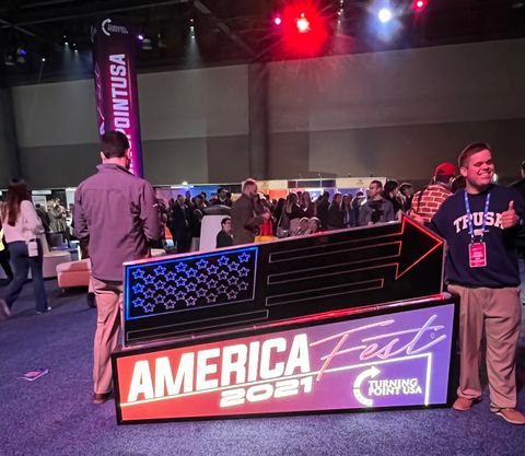 A young man in a TPUSA sweater poses next to a neon AmericaFest 2021 sign in the exhibitor hall. A crowded group of people walk around the hall in the background, including a man in a brick pattern suit. The room is lit up with red, white, and blue