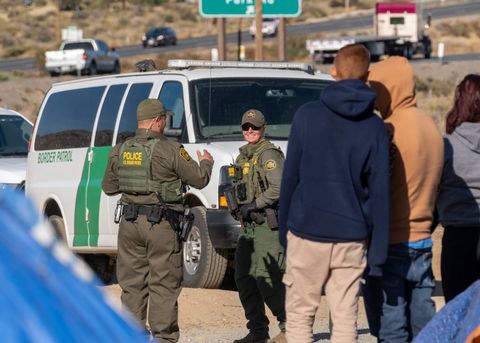 In the foreground, the edges of tents are visible but out of focus. Beyond those tents are asylum seekers, who are standing with their backs to the camera. Beyond them, a border patrol agent is smiling at another agent, who is gesticulating and facing away from the camera. Behind the agents is a border patrol van, and in the distance one can see a highway.