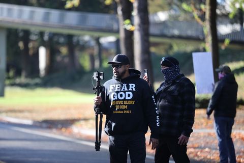 Black Conservative Preacher wearing a 'FEAR GOD And Give Glory to Him' black sweatshirt. He's standing in the street holding up a tripod with a cell-phone attached at the top, livestreaming the crowd across the street. Behind him, another individual, but with their face covered, is also recording via a cellphone.