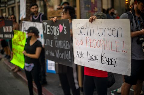 A woman holds a sign that says 'SETH GREEN IS A PEDOPHILE!..ASK ISAAC KAPPIE.'