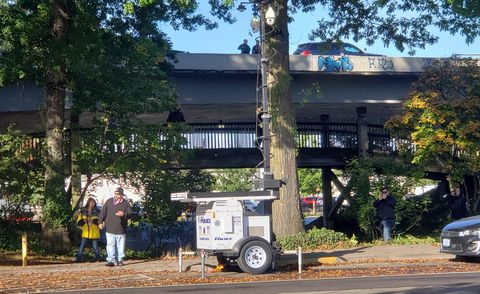 A photograph looking towards the Washington-Jefferson skate park in Eugene. In the direct middle is white-painted two-wheeled trailer propped up with four legs. Expanding upwards is a black pole with two cameras attached at top. In the background, and around, are a handful of bigots both on the ground and on a curved bridge behind the camera. Even further, and higher, is the I-105 overpass where two EPD officers can be seen standing in-between two cruisers.