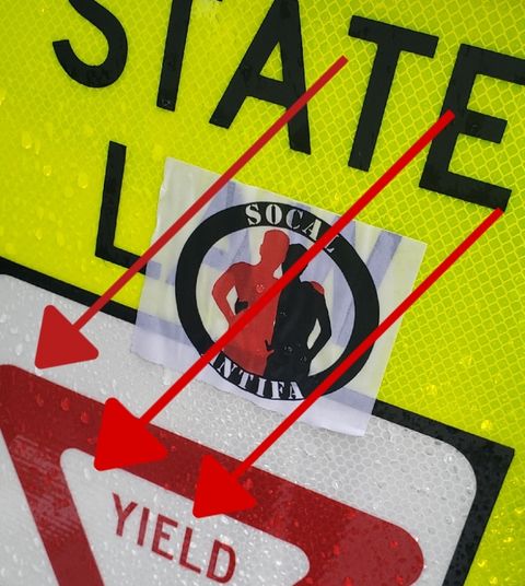A sticker on a traffic sign labeled socal antifa which depicts silhouettes of two people rubbing their penises on each other