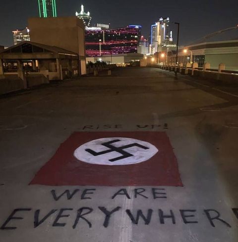 Neo-Nazi graffiti of a swastika with the caption at the top 'rise up' and the caption at the bottom 'we are everywhere' on the roof of a parking garage with the downtown Dallas skyline at night in the background.