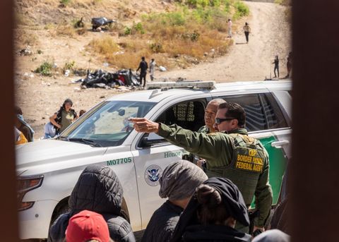 a border agent points to something off screen in front of a crowd of asylum seekers