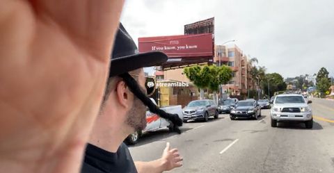 a man wearing a mocking imitation of a hasidic jewish man's dress holds his hand on a camera and looks out at traffic as he stands in the street