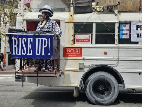 The back of a white school bus with a DJ in drag with a banner over her DJ both taht says "rise up!"