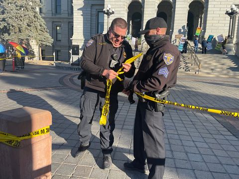 Two Colorado state patrol officers attempt to put tape back up knocked over by the wind. About 50 feet behind them there are WBC members holding various signs with homophobic and antisemitic messages