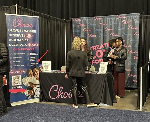  a table with a black tarp reading “Choices” in pink text sits in a hall lined with black drapes. Behind their table is a patterned banner reading “Choices” all over. Four women stand around the booth talking. A banner to the side reads “Choices because women deserve better and babies deserve a chance.” The banner displays a list of services including “abortion pill reversal.”