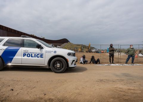 Four protesters block the fenced resource site. Two are sitting, two are standing. To the left of the frame, a Federal Protective Service vehicle is entering, driving to the right of the frame. It will pass between the protesters and the camera. The border wall is seen as it stretches into the distance.