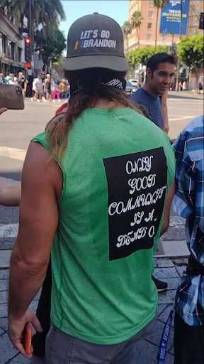 A man in a green sleeveless shirt stands with his back to the camera. The back of his shirt reads “The only good communist is a dead one” and he is wearing a hat that says “Let’s Go Brandon.”