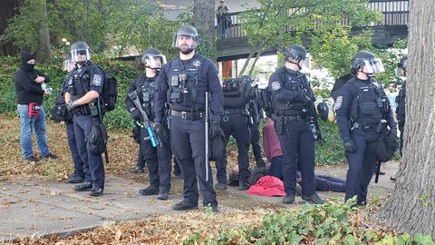 At least seven police officers in riot gear form a semi-circle around a man lying on the ground behind them. One officer is holding a less-lethal munition launcher.